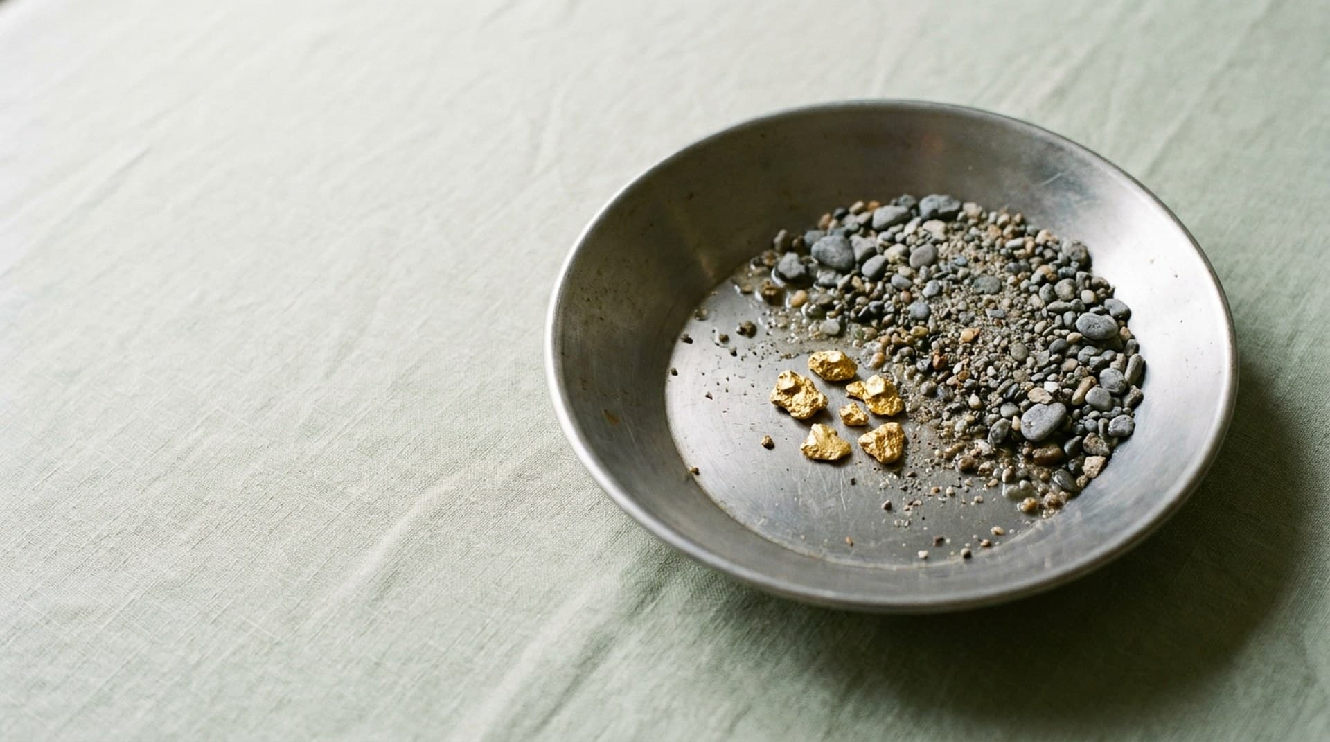 Meta conversion leads optimization: separating qualified leads from form fills A steel gold prospector's pan on sage linen with a few gold nuggets separated from grey river gravel, representing Meta conversion leads optimization filtering qualified leads from form fills