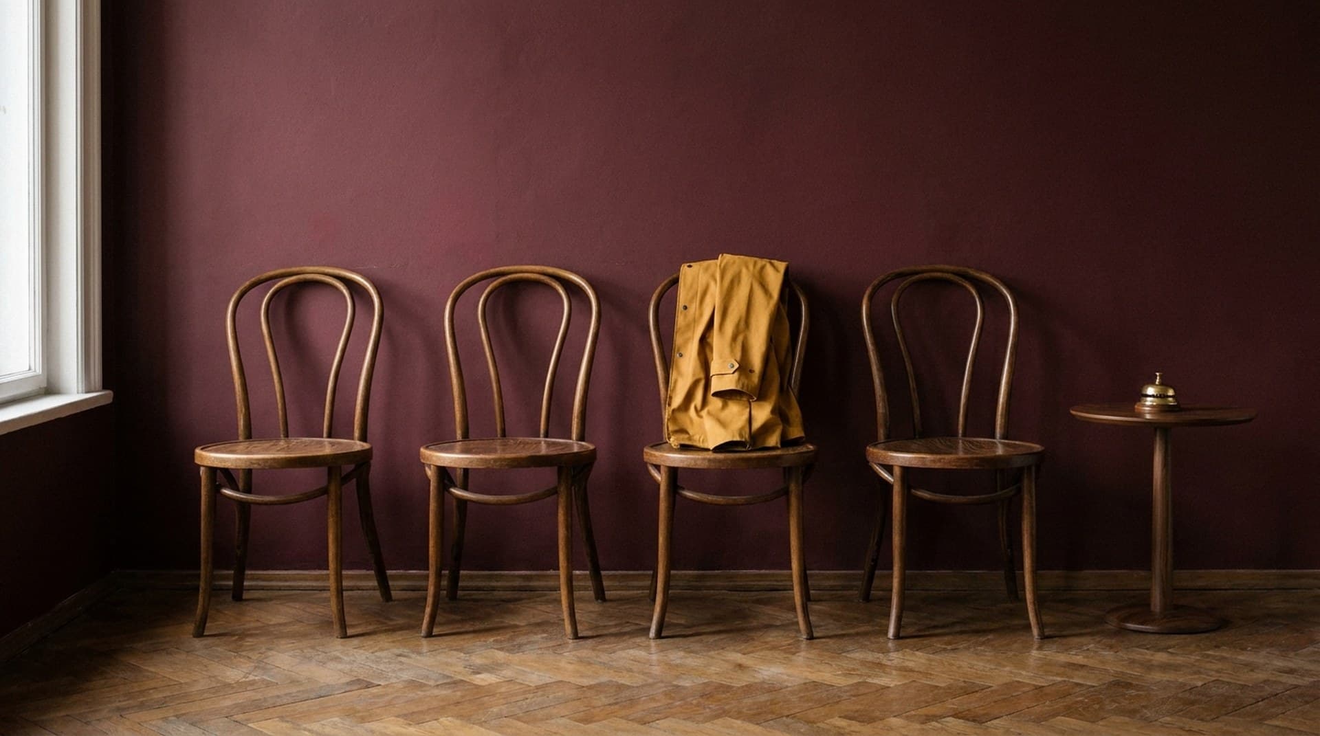 A waiting execution only moves when someone rings the right bell. Four vintage bentwood chairs against a burgundy wall in an empty waiting room, one holding a folded mustard raincoat, with a brass service bell on a side table, a metaphor for an n8n Wait node waiting for a resume webhook call